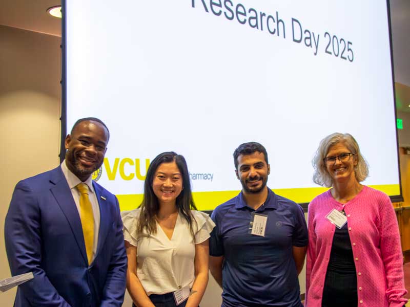 Dean K.C. Ogbonna and Associate Dean Mary Peace McRae stand on either side of graduate student poster award winners Grace Rilee and Yasir Alshehry, at the front of a lecture hall on Research Day.
